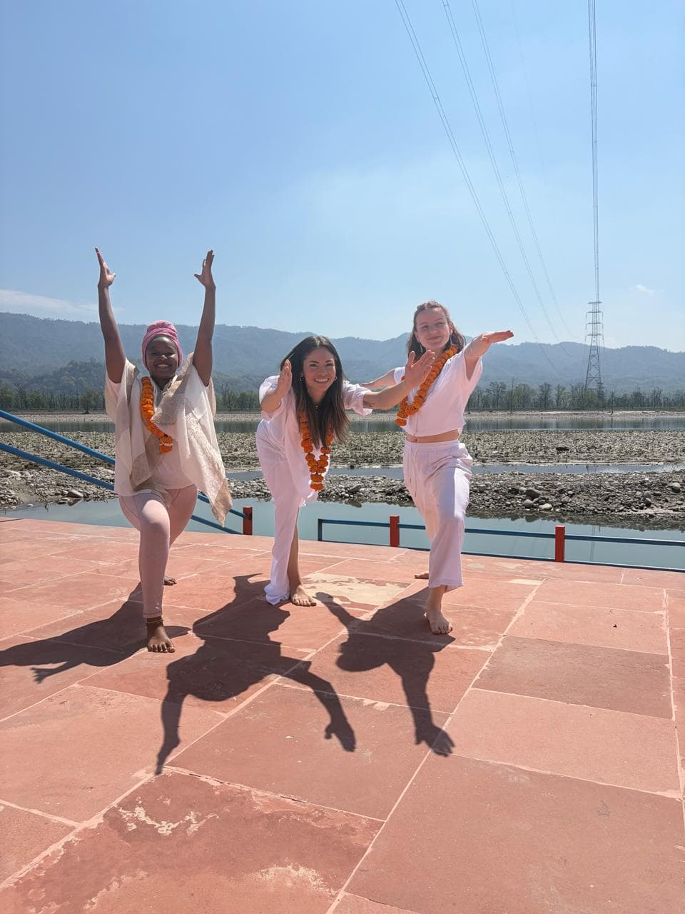 Three women practicing yoga outdoors near a river with mountains in the background.